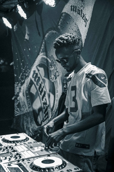 A person wearing a sports jersey operates a DJ mixing console in a dimly lit environment. The background features a banner with abstract designs, and stage lights illuminate part of the scene, creating a focused and moody ambiance.