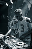 A person wearing sunglasses and a sports jersey is focused on operating DJ equipment. The scene is in a dimly lit area with a backdrop that seems to have the image of a beverage bottle and some text.