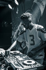 A person wearing sunglasses and a sports jersey is focused on operating DJ equipment. The scene is in a dimly lit area with a backdrop that seems to have the image of a beverage bottle and some text.