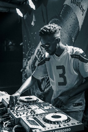 A person wearing sunglasses and a sports jersey is focused on operating DJ equipment. The scene is in a dimly lit area with a backdrop that seems to have the image of a beverage bottle and some text.