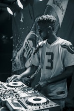 A person wearing sunglasses and a football jersey operates a DJ booth. The lighting is dim and atmospheric, with a large banner displaying a brand name in the background.