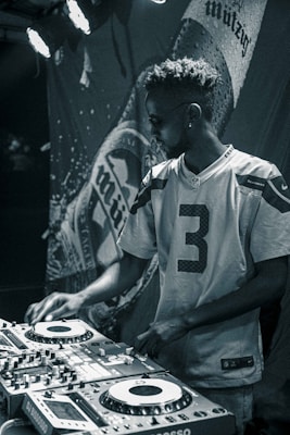 A person wearing sunglasses and a football jersey operates a DJ booth. The lighting is dim and atmospheric, with a large banner displaying a brand name in the background.