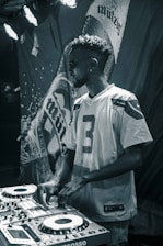 A man wearing sunglasses and a sports jersey is operating DJ equipment with a focused expression. The background features a large, textured banner with partially visible text. Stage lights illuminate the scene with a moody atmosphere.