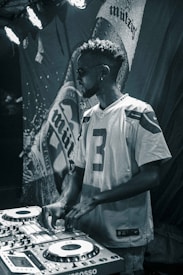 A man wearing sunglasses and a sports jersey is operating DJ equipment with a focused expression. The background features a large, textured banner with partially visible text. Stage lights illuminate the scene with a moody atmosphere.
