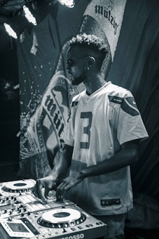 A man wearing sunglasses and a sports jersey is operating DJ equipment with a focused expression. The background features a large, textured banner with partially visible text. Stage lights illuminate the scene with a moody atmosphere.