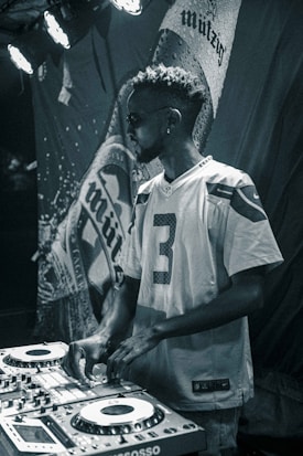 A man wearing sunglasses and a sports jersey is operating DJ equipment with a focused expression. The background features a large, textured banner with partially visible text. Stage lights illuminate the scene with a moody atmosphere.