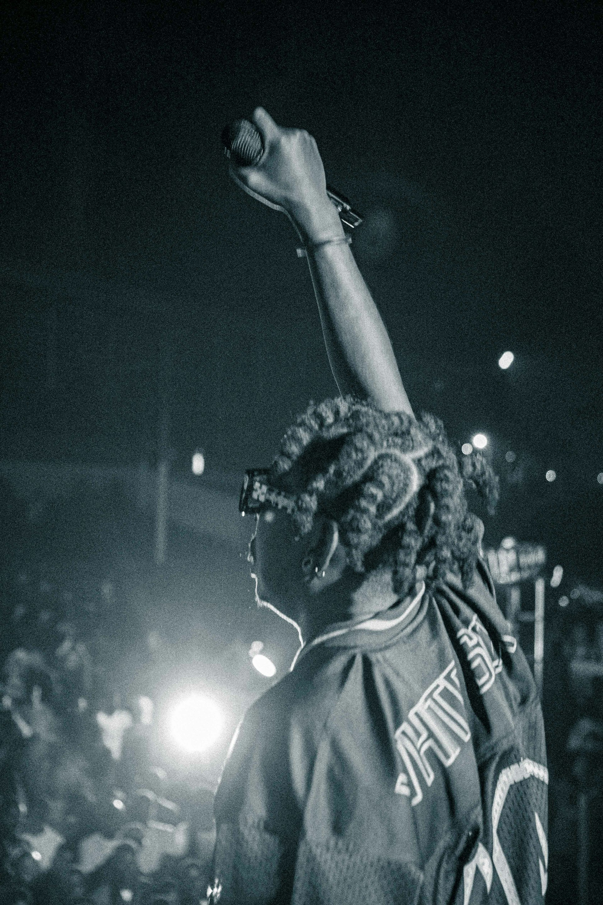 A performer with braided hair is on stage, holding a microphone up with one hand. The lighting is dim, with bright stage lights in the background, casting a silhouette effect. The performer is wearing a jersey, and the audience can be seen faintly in front.