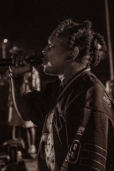 A person with dreadlocks is captured mid-performance, holding a microphone close to their mouth. They are wearing a sports jersey with visible numbers and lettering. The background is dimly lit, creating a moody atmosphere with blurred figures.