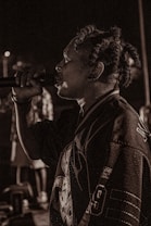 A person with dreadlocks is captured mid-performance, holding a microphone close to their mouth. They are wearing a sports jersey with visible numbers and lettering. The background is dimly lit, creating a moody atmosphere with blurred figures.