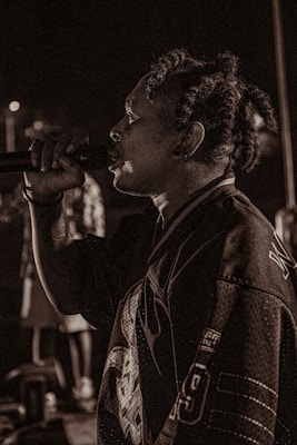 A person with dreadlocks is captured mid-performance, holding a microphone close to their mouth. They are wearing a sports jersey with visible numbers and lettering. The background is dimly lit, creating a moody atmosphere with blurred figures.