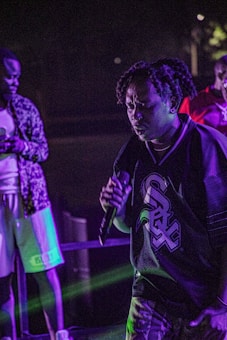 A person holding a microphone is performing on stage, wearing a sports jersey with bold typography. The setting appears to be a nightlife scene with purple lighting and other people nearby.