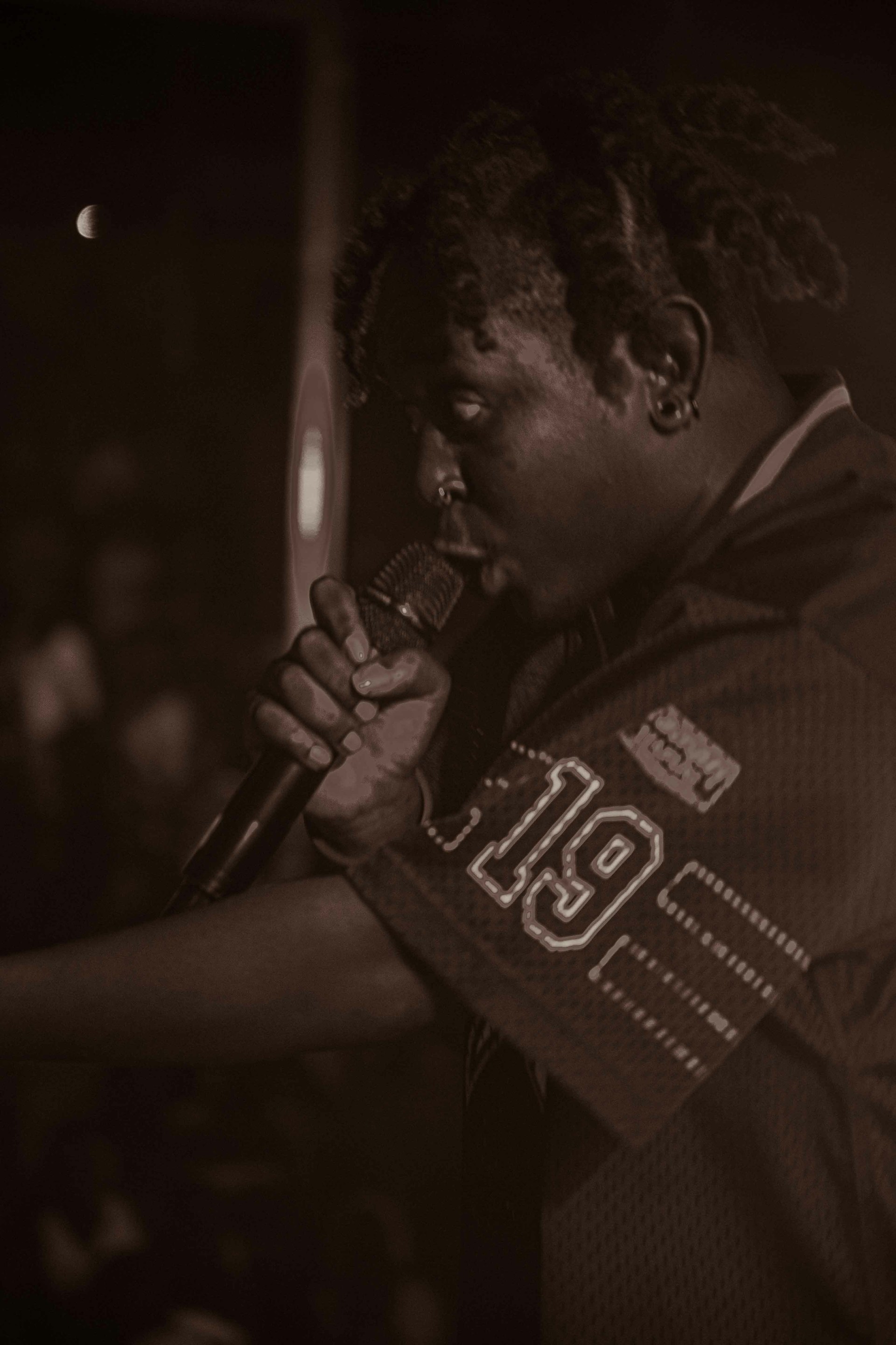 A person with braided hair is passionately performing with a microphone. The setting is dimly lit, possibly indicating a concert or music event. They are wearing a jersey with the number 19 on it. The background is blurred, highlighting the focus on the performer.