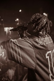 Captured in a dimly lit setting, a performer holds a microphone, engaging with the audience. The crowd is partially visible, with blurred lights in the background creating a lively atmosphere. The performer wears a jersey with visible text on the back.
