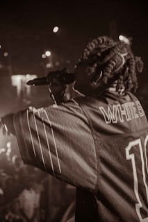 Captured in a dimly lit setting, a performer holds a microphone, engaging with the audience. The crowd is partially visible, with blurred lights in the background creating a lively atmosphere. The performer wears a jersey with visible text on the back.