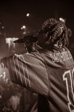 Captured in a dimly lit setting, a performer holds a microphone, engaging with the audience. The crowd is partially visible, with blurred lights in the background creating a lively atmosphere. The performer wears a jersey with visible text on the back.