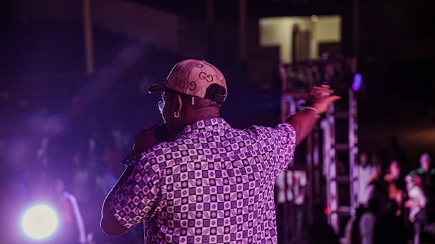 A person wearing a patterned shirt and a cap, holding a microphone, is on stage performing or speaking to an audience. The lighting is dim with a spotlight illuminating the scene.