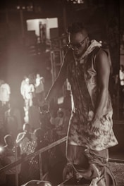 A person in stylish attire and sunglasses is performing on stage in front of an audience. Stage lighting creates a dramatic atmosphere with a mix of light and shadow.