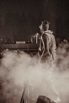 A person stands on a stage or platform enveloped in thick smoke or fog, creating a mysterious and atmospheric setting. The individual is wearing a sleeveless jacket and sunglasses, with their hair styled in an updo. The background is dark, with minimal lighting that highlights the smoke.