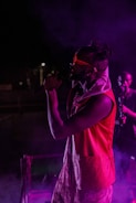 A performer on stage wearing a vibrant red vest and sunglasses, illuminated by purple and pink stage lights, passionately singing into a microphone. Smoke effects add a dramatic atmosphere, with another person visible in the background.