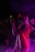 A performer on stage wearing a vibrant red vest and sunglasses, illuminated by purple and pink stage lights, passionately singing into a microphone. Smoke effects add a dramatic atmosphere, with another person visible in the background.
