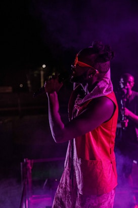A performer on stage wearing a vibrant red vest and sunglasses, illuminated by purple and pink stage lights, passionately singing into a microphone. Smoke effects add a dramatic atmosphere, with another person visible in the background.