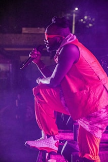 A performer wearing bright orange clothing takes a dynamic stance on stage while holding a microphone. The individual has stylish sunglasses and is lit by purple stage lighting, creating an energetic atmosphere.