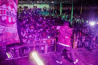 A performer on stage faces a large, enthusiastic crowd. Bright lighting and vibrant colors create a lively atmosphere. A prominent advertisement for a brand of beer is visible on the side.
