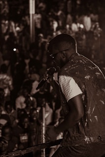 A performer holds a microphone on stage in front of a large, crowded audience. The scene is captured in a monochromatic color scheme with the focus on the singer and the audience members in the background.
