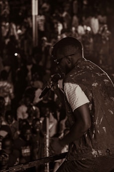 A performer holds a microphone on stage in front of a large, crowded audience. The scene is captured in a monochromatic color scheme with the focus on the singer and the audience members in the background.