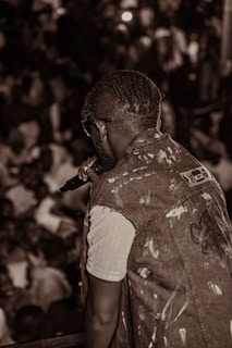 A person with braided hair and sunglasses is holding a microphone, facing a large, crowded audience. They are wearing a light-colored shirt underneath a sleeveless denim vest. The scene is lively and captured in sepia tones.