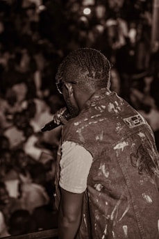 A person with braided hair and sunglasses is holding a microphone, facing a large, crowded audience. They are wearing a light-colored shirt underneath a sleeveless denim vest. The scene is lively and captured in sepia tones.