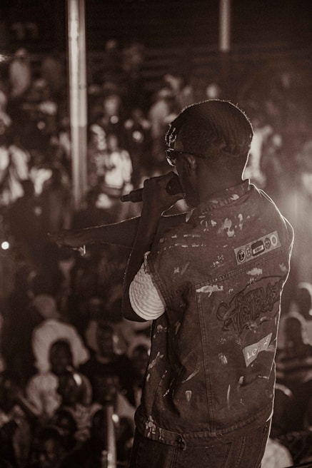 A singer on stage is holding a microphone, performing in front of a large crowd. The scene is captured in sepia tones, giving it a classic and nostalgic feel. The backdrop is a blurred audience, indicating movement and energy.
