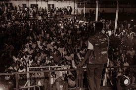 A person stands on a stage facing a large, tightly packed audience in a dimly lit venue. The crowd is diverse and energetic, with individuals holding up phones with flashlights on to capture the moment. The person on stage is wearing a denim vest with various patches, including one that says 'Levi's'. The atmosphere is vibrant and anticipatory, reflecting a concert or public event.