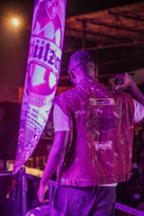 A person with braided hair, wearing a Levi's denim jacket, is standing on a stage or platform holding a microphone. There is a large, illuminated advertisement or banner with the word 'Hürzog' behind them, adding a prominent purple hue to the scene.