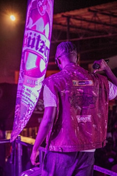 A person with braided hair, wearing a Levi's denim jacket, is standing on a stage or platform holding a microphone. There is a large, illuminated advertisement or banner with the word 'Hürzog' behind them, adding a prominent purple hue to the scene.