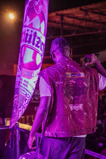 A person with braided hair, wearing a Levi's denim jacket, is standing on a stage or platform holding a microphone. There is a large, illuminated advertisement or banner with the word 'Hürzog' behind them, adding a prominent purple hue to the scene.