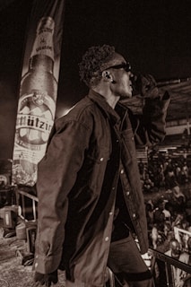 A person wearing sunglasses and a dark jacket is holding a microphone on stage. In the background, there is a large banner displaying a beer bottle with the label 'Mützig'. The surrounding area is crowded with people, suggesting a concert or live event. The image has a sepia tone, adding a vintage feel.