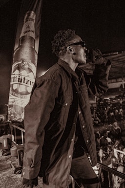 A person wearing sunglasses and a dark jacket is holding a microphone on stage. In the background, there is a large banner displaying a beer bottle with the label 'Mützig'. The surrounding area is crowded with people, suggesting a concert or live event. The image has a sepia tone, adding a vintage feel.