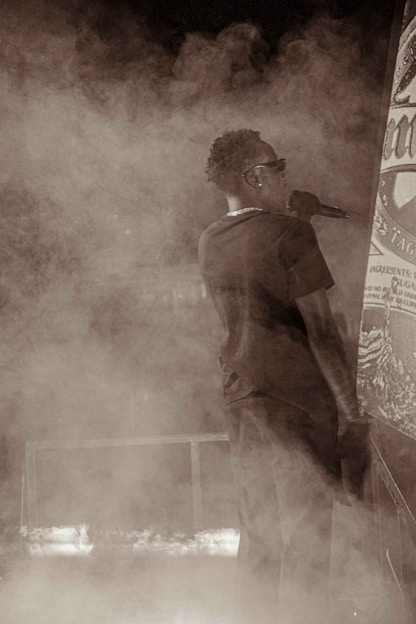 A person in a dark outfit holds a microphone while standing amidst swirling smoke or fog. They are facing sideways, wearing sunglasses, and appear to be on a stage with a large poster or banner nearby.