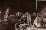 A performer with a microphone appears to be passionately engaging an audience onstage, accompanied by another individual in a less illuminated part of the stage. Advertising banners and dimly lit surroundings suggest a lively outdoor concert environment. Smoke or fog enhances the ambiance.