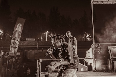 A performer with a microphone appears to be passionately engaging an audience onstage, accompanied by another individual in a less illuminated part of the stage. Advertising banners and dimly lit surroundings suggest a lively outdoor concert environment. Smoke or fog enhances the ambiance.