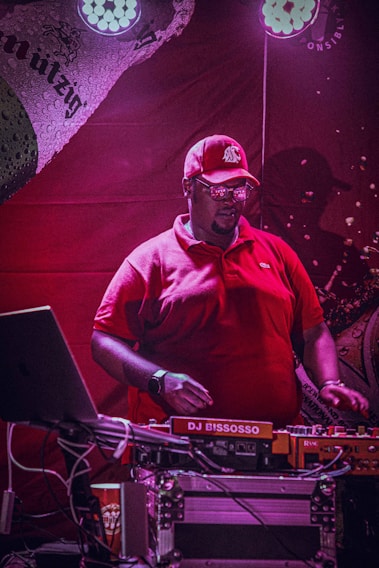 A person wearing a red cap and shirt is standing behind a DJ setup with equipment labeled DJ BISSOSSO. The background features red lighting, creating a vibrant atmosphere, and spotlights illuminate the scene.