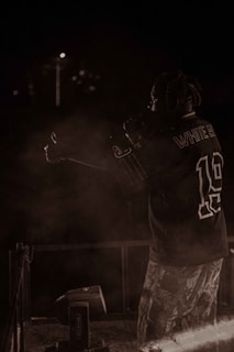 A person with braided hair is on stage, holding a microphone, wearing a sports jersey and patterned pants. The background is dark, with minimal lighting and some smoke or haze around the performer.