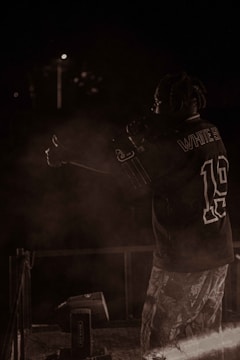 A person with braided hair is on stage, holding a microphone, wearing a sports jersey and patterned pants. The background is dark, with minimal lighting and some smoke or haze around the performer.