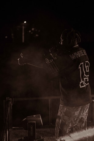 A person with braided hair is on stage, holding a microphone, wearing a sports jersey and patterned pants. The background is dark, with minimal lighting and some smoke or haze around the performer.
