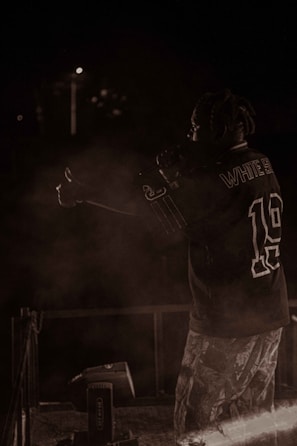 A person with braided hair is on stage, holding a microphone, wearing a sports jersey and patterned pants. The background is dark, with minimal lighting and some smoke or haze around the performer.