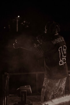 A person with braided hair is on stage, holding a microphone, wearing a sports jersey and patterned pants. The background is dark, with minimal lighting and some smoke or haze around the performer.