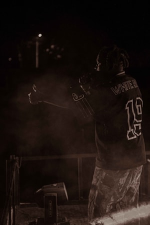 A person with braided hair is on stage, holding a microphone, wearing a sports jersey and patterned pants. The background is dark, with minimal lighting and some smoke or haze around the performer.