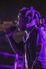 A close-up of a person speaking softly into a sleek microphone, bathed in deep blue and amber light.