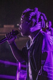 A person holding a microphone stands in profile under dramatic lighting, with purple hues illuminating the scene. The background is dark, highlighting the individual's presence and attire.